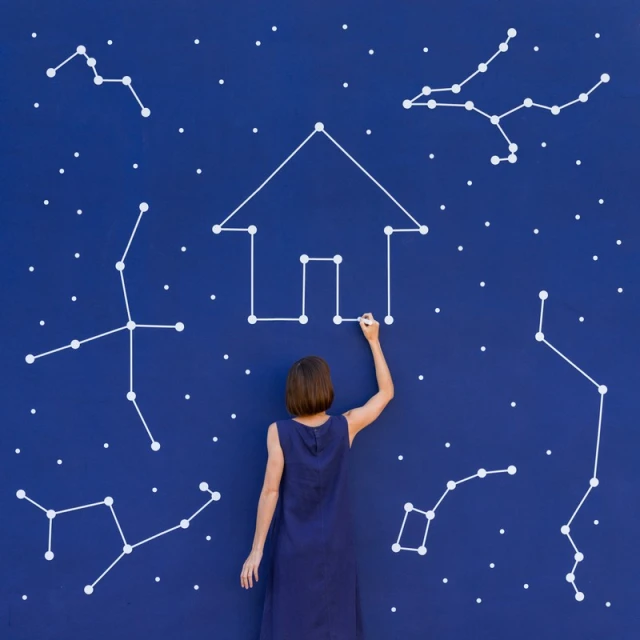 Back view of a woman drawing a house with constellations on a blue wall, creating a whimsical, celestial real estate concept.