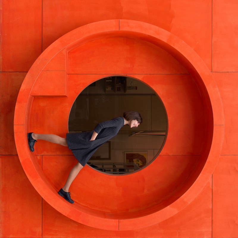 Woman posing inside a large orange circular sculpture against a matching wall with a peek into another space through a smaller circle.
