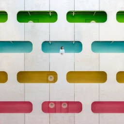 A person standing in an oval cutout of a geometric concrete facade featuring colorful recessed window rows.