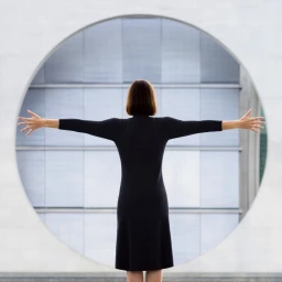 Woman in a black dress standing with arms outstretched in front of a large circular architectural window opening.