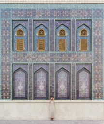 A woman in a cream-colored dress standing against an ornate blue tiled wall with arched windows, photographed by Anna Devís and Daniel Rueda.