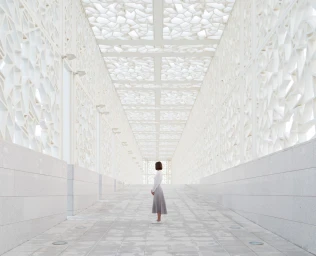 Woman in white top and grey pleated skirt standing inside a white geometric architectural walkway with patterned ceiling