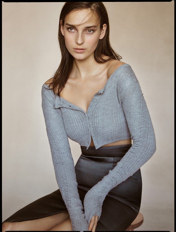 Portrait of a young woman with long brown hair wearing a knitted crop top and a pencil skirt, posing against a neutral background.