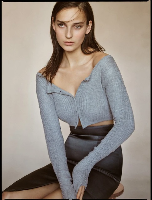 Portrait of a young woman with long brown hair wearing a knitted crop top and a pencil skirt, posing against a neutral background.