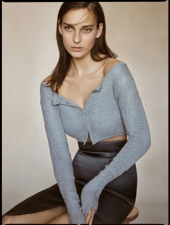 Portrait of a young woman with long brown hair wearing a knitted crop top and a pencil skirt, posing against a neutral background.