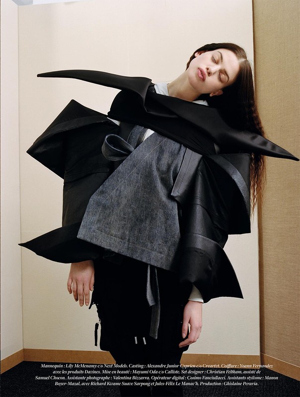 A fashion-forward photo captures a female model in an avant-garde black outfit with an unusual shape, posed indoors against a neutral background.