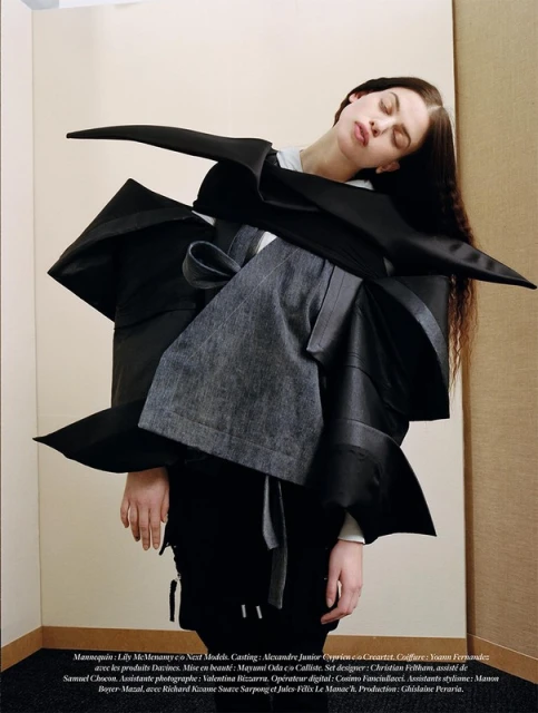 A fashion-forward photo captures a female model in an avant-garde black outfit with an unusual shape, posed indoors against a neutral background.