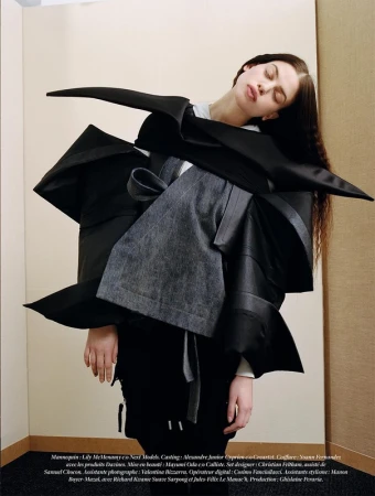 A fashion-forward photo captures a female model in an avant-garde black outfit with an unusual shape, posed indoors against a neutral background.