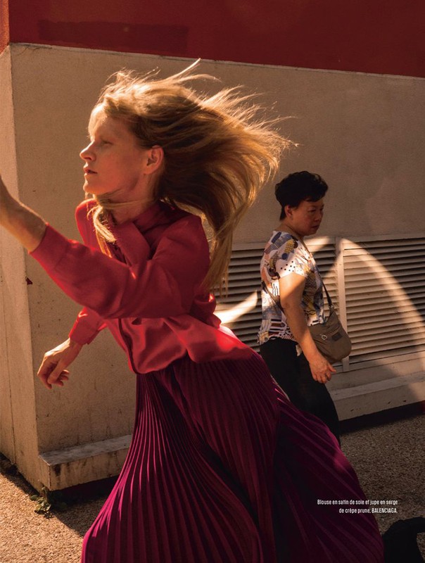 Woman in flowing purple skirt and red top walks past another woman in an urban setting, with shadows and sunlight creating contrast.