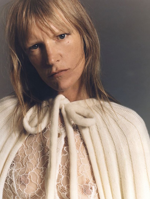Close-up of a blonde adult woman with a neutral expression, wearing a knitted cape and lace top in a studio setting. Fashion portrait.