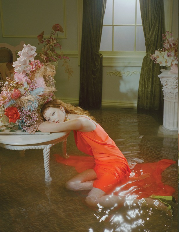 Woman in orange dress poses in a room with water-covered floors, flowers on a table, and light coming through a window.