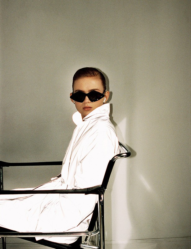 A fair-skinned person with short brown hair, wearing sunglasses and a shiny white jacket, is seated in a black chair against a light grey wall.