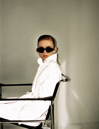 A fair-skinned person with short brown hair, wearing sunglasses and a shiny white jacket, is seated in a black chair against a light grey wall.
