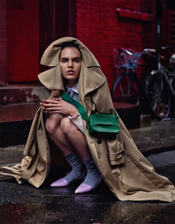 Model in oversized beige trench coat hooded over head wearing purple heels and a green crossbody bag