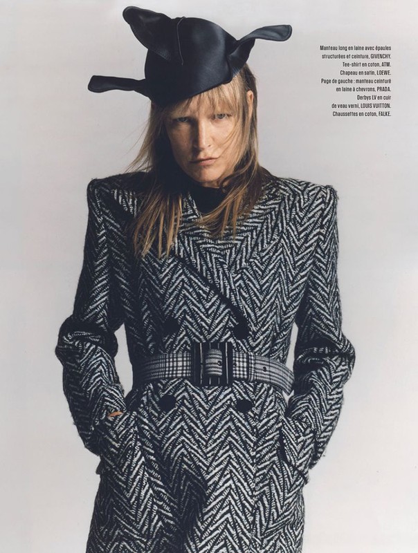 Blonde fashion model poses in a patterned black and white coat with large black belt, and matching hat against a neutral backdrop.