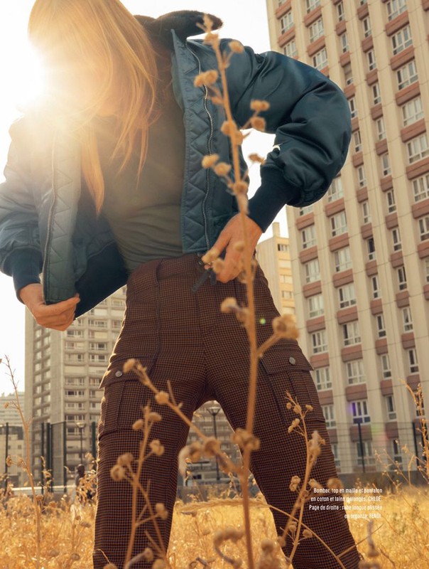 Woman in bomber jacket and patterned pants stands in a field with tall buildings in background, bathed in warm sunlight.