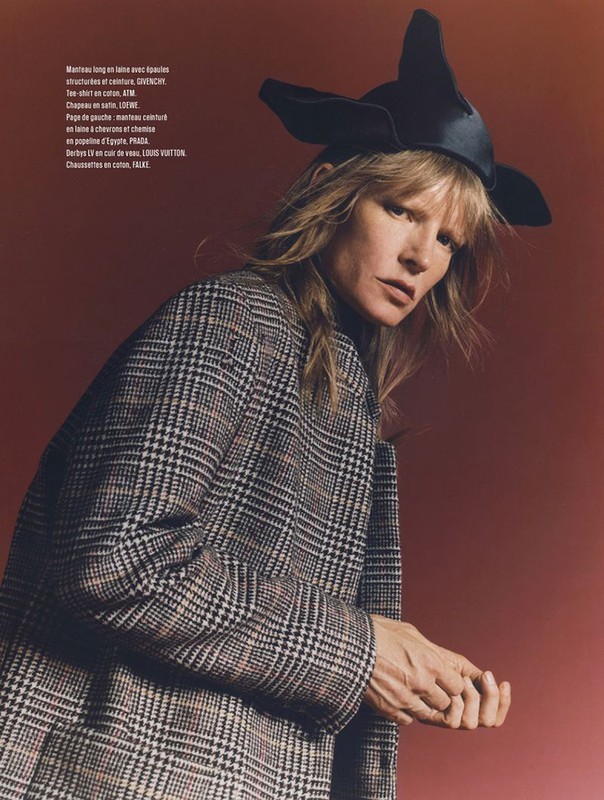 Fashion editorial shot of a fair-skinned woman wearing a plaid coat and sculptural hat against a reddish-brown backdrop.
