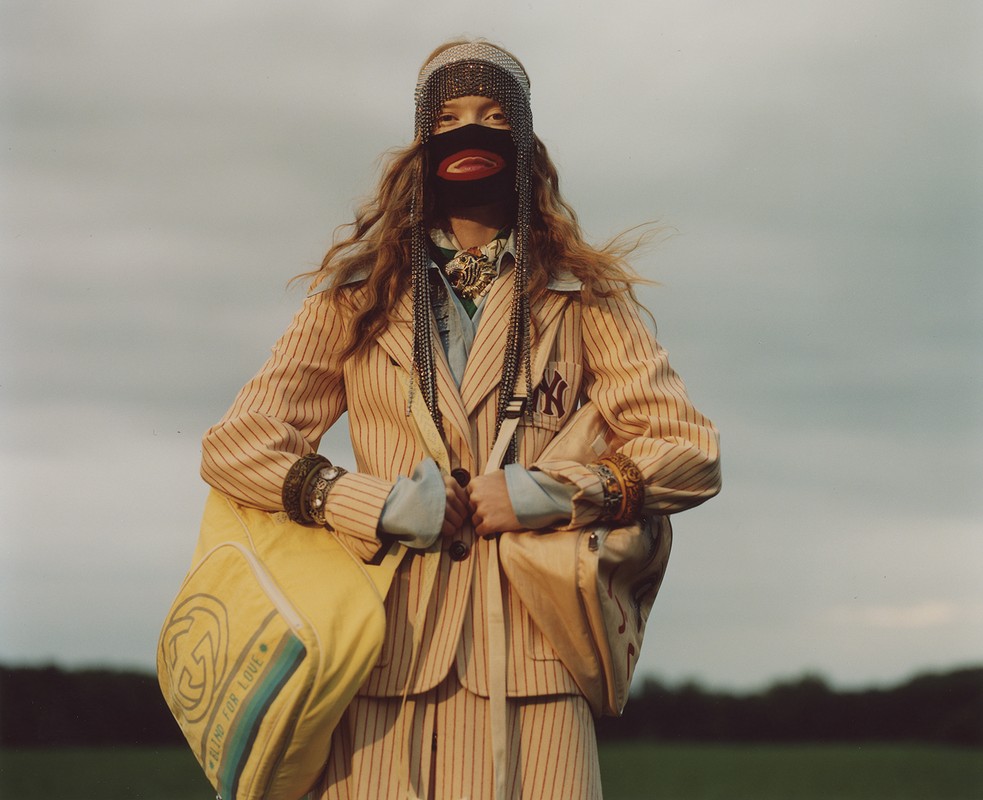 Fashion model in pinstripe suit, face mask, and chainmail headpiece holds a yellow duffel bag in an outdoor field setting.