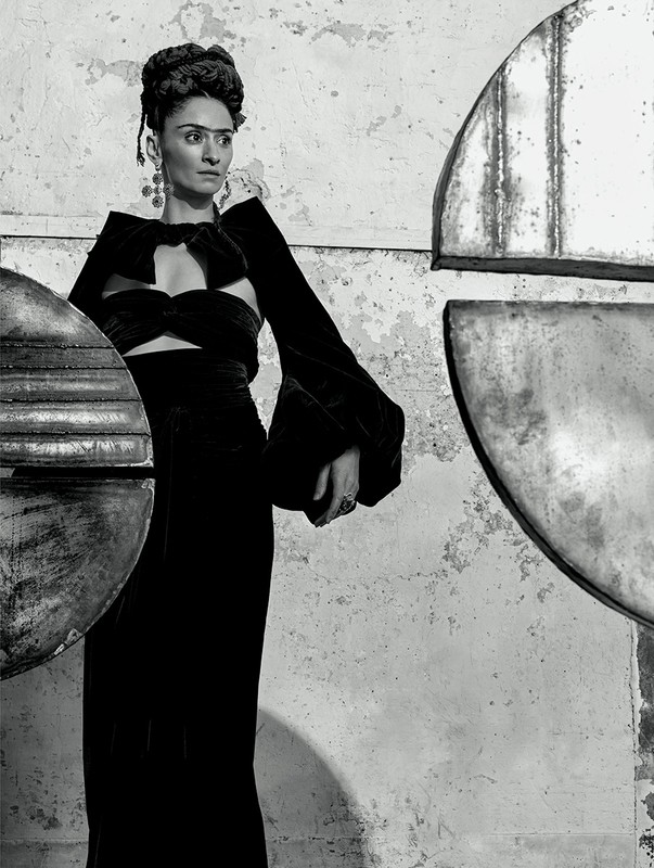 Monochrome portrait of a woman in a cut-out velvet dress with hair up, dramatic earrings, against a textured background with metal shapes.