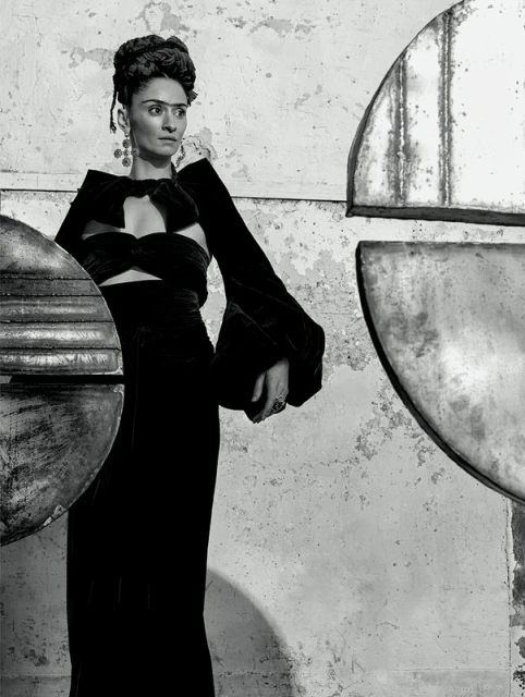 Monochrome portrait of a woman in a cut-out velvet dress with hair up, dramatic earrings, against a textured background with metal shapes.