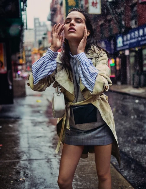 A woman in a beige trench coat stands in the rain in a city street. She wears a striped shirt and a gray skirt.