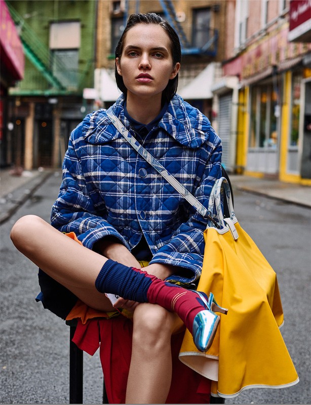 Woman with short dark hair sits in an urban alleyway wearing a blue quilted jacket, orange skirt, and blue metallic high heels.