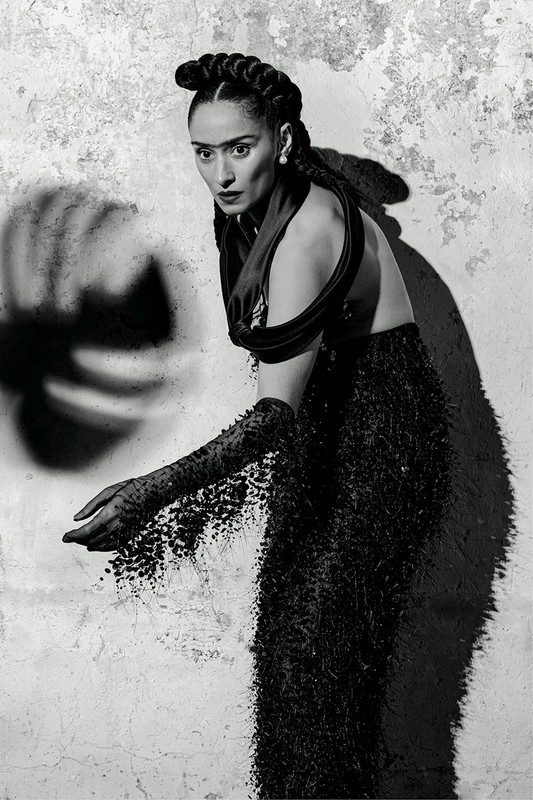 A black-and-white portrait of a woman in dark formalwear with artistic makeup, posed against a textured wall with shadow effects.