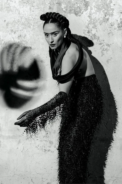 A black-and-white portrait of a woman in dark formalwear with artistic makeup, posed against a textured wall with shadow effects.