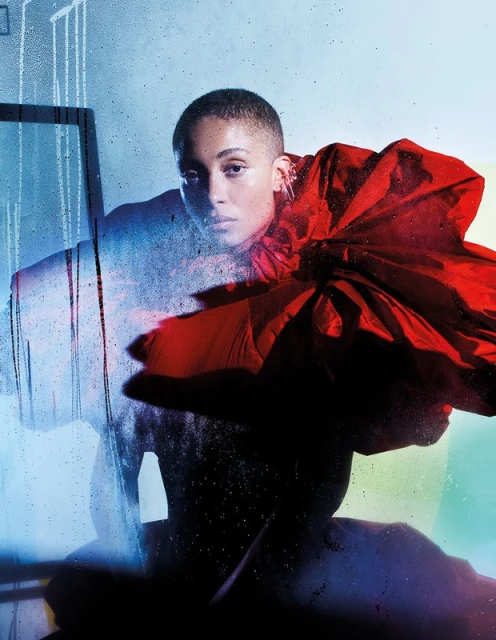 Headshot of a bald woman with fair skin in an oversized red flower-like garment, posing in front of a blue-toned background.