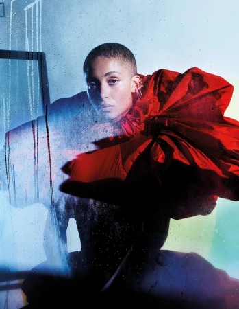Headshot of a bald woman with fair skin in an oversized red flower-like garment, posing in front of a blue-toned background.