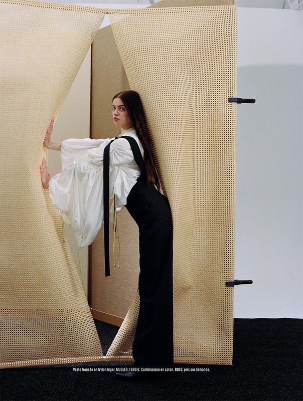 A young adult woman with long brown hair, wearing a white blouse and black overalls, poses in front of a tan and white woven backdrop.