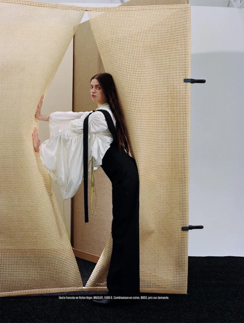 A young adult woman with long brown hair, wearing a white blouse and black overalls, poses in front of a tan and white woven backdrop.