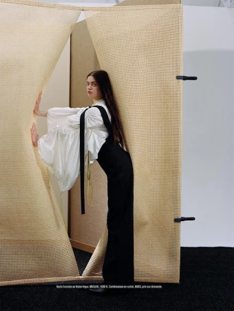 A young adult woman with long brown hair, wearing a white blouse and black overalls, poses in front of a tan and white woven backdrop.