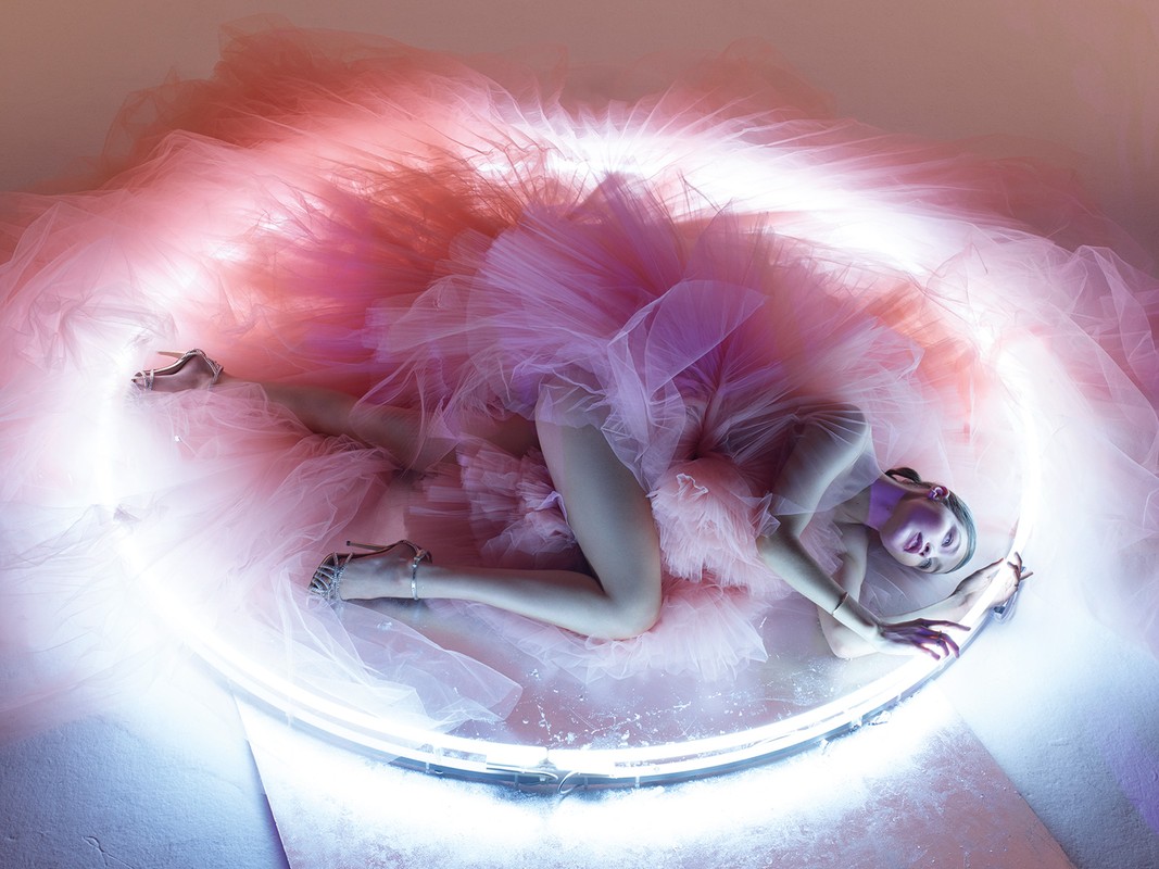 Woman in pink tulle gown lying next to illuminated ring, studio shot. Fashion, beauty, and artistic concept with pink and white tones.