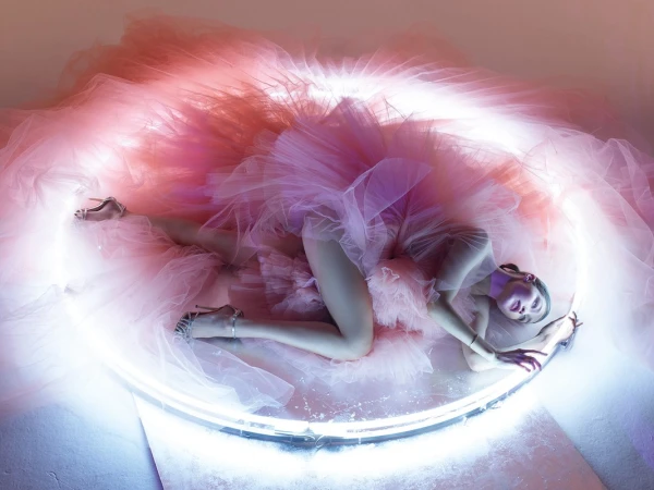Woman in pink tulle gown lying next to illuminated ring, studio shot. Fashion, beauty, and artistic concept with pink and white tones.