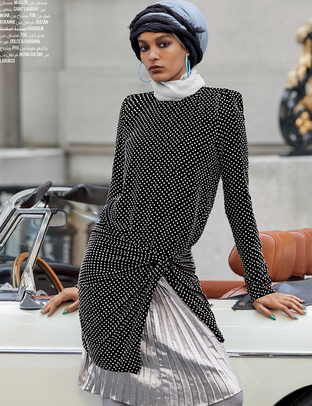 Fashion editorial shot of a female model wearing a patterned top, pleated skirt, and head wrap, posing outdoors with a white car.