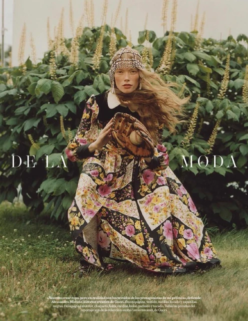 A woman in a floral dress poses outdoors holding a baseball glove, set against a green leafy backdrop, in a fashion editorial shot.