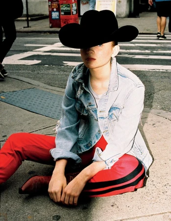 Young adult sitting on a city street wearing a cowboy hat, denim jacket, and red pants with black stripes, street scene in the background.