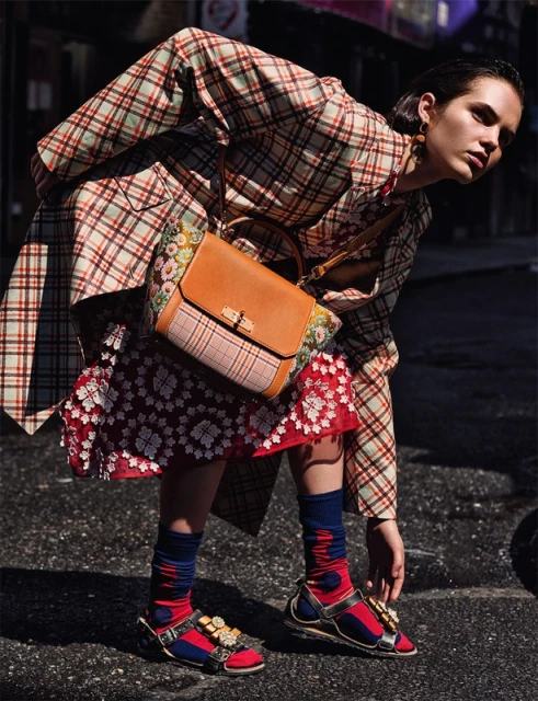 A young adult woman poses in a fashion editorial shot, wearing a plaid trench coat, red floral skirt, and embellished sandals on a city street.