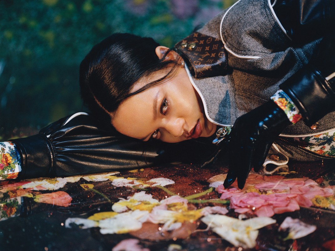 Close-up portrait of a woman with dark hair, wearing patterned jacket and gloves, lying on a surface with pink and white flowers.