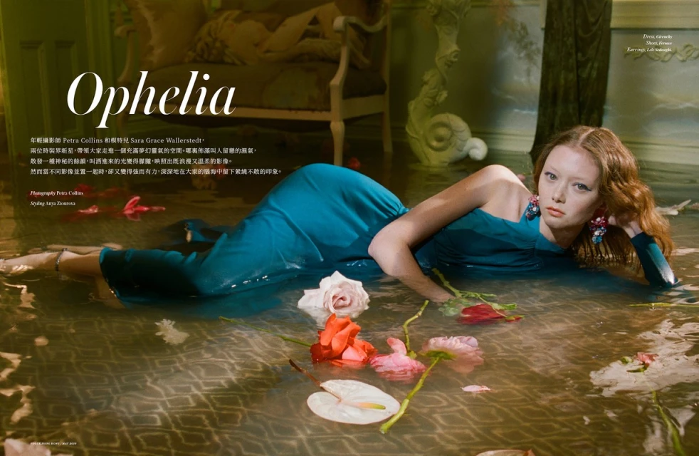 Red-haired woman in a teal dress poses in shallow water surrounded by flowers, illuminated by soft light in an indoor setting with ornate decor.