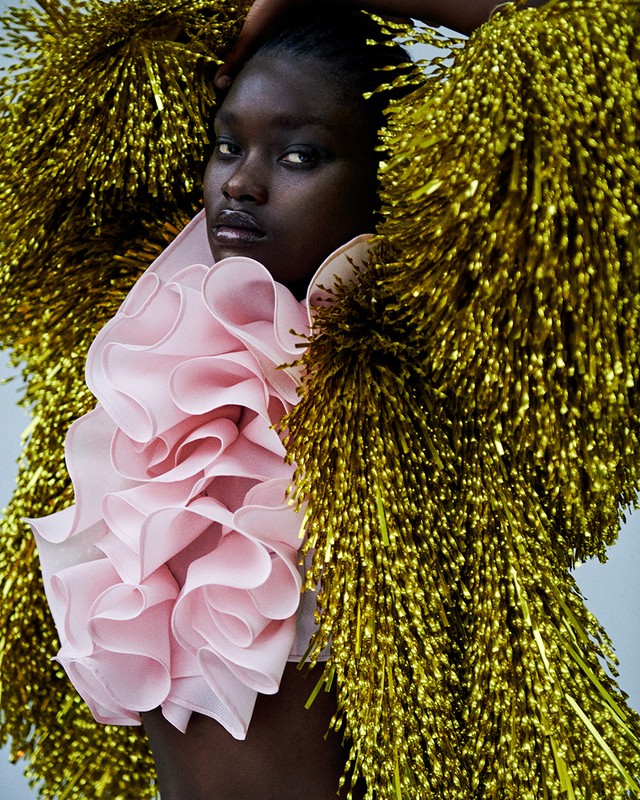 Close-up of a Black female model wearing a pink ruffled top and gold tinsel jacket in a beauty and fashion editorial shot.