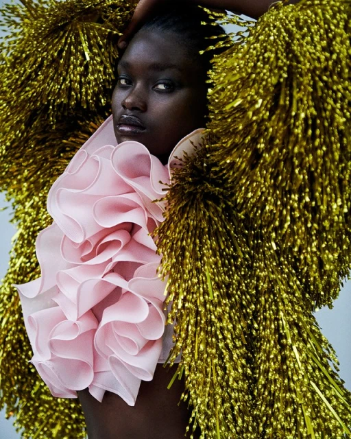 Close-up of a Black female model wearing a pink ruffled top and gold tinsel jacket in a beauty and fashion editorial shot.