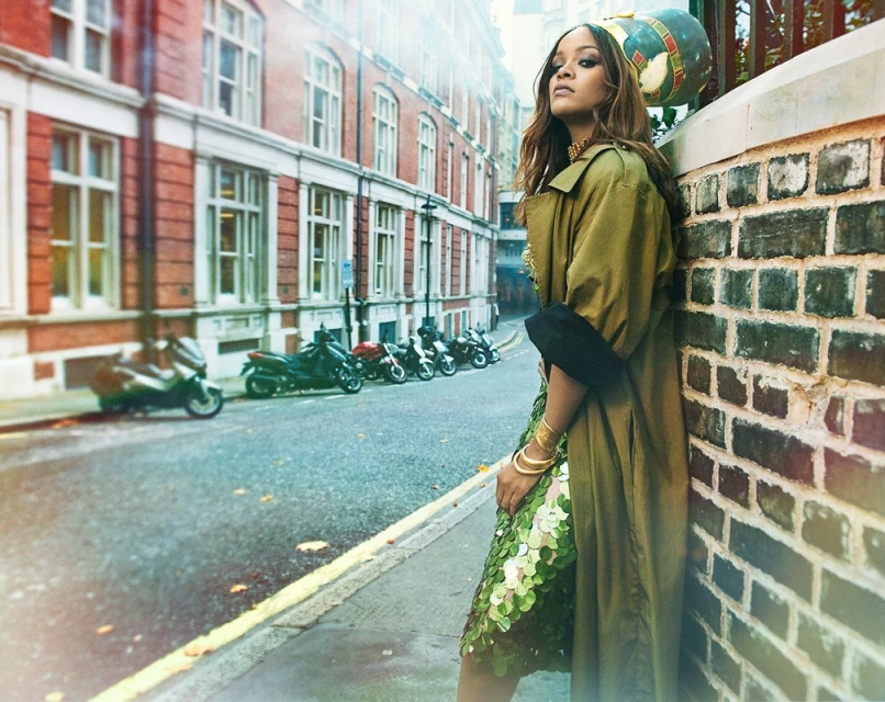 Brunette woman in trench coat and green sequined dress poses against brick wall on urban street with scooters and buildings.