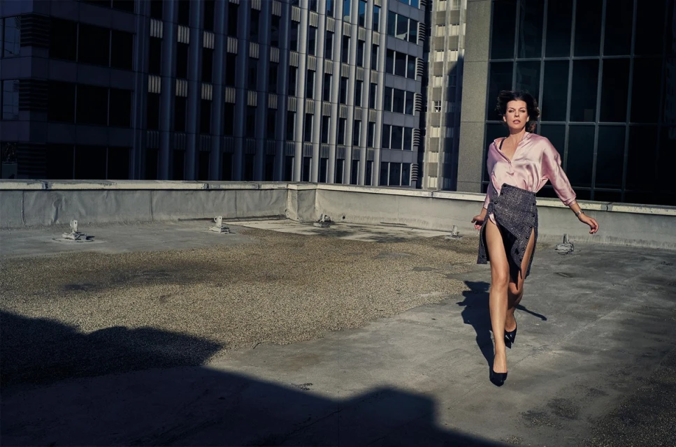 Fashion shot of an adult woman in a pink blouse and dark skirt walking on a rooftop with a tall building in the background.