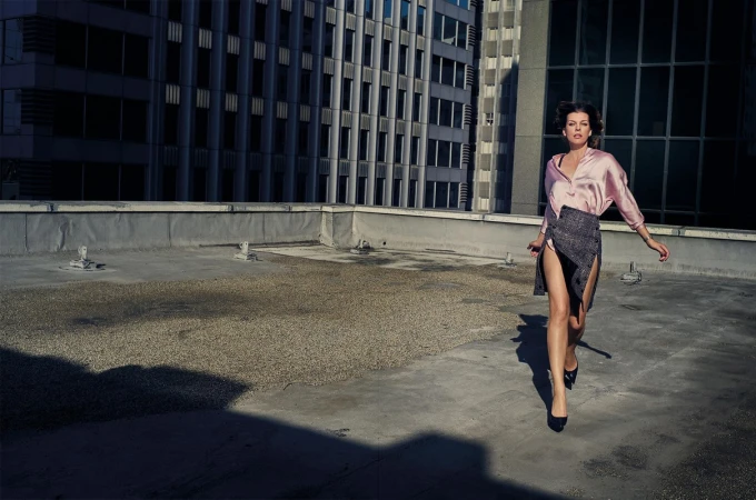 Fashion shot of an adult woman in a pink blouse and dark skirt walking on a rooftop with a tall building in the background.
