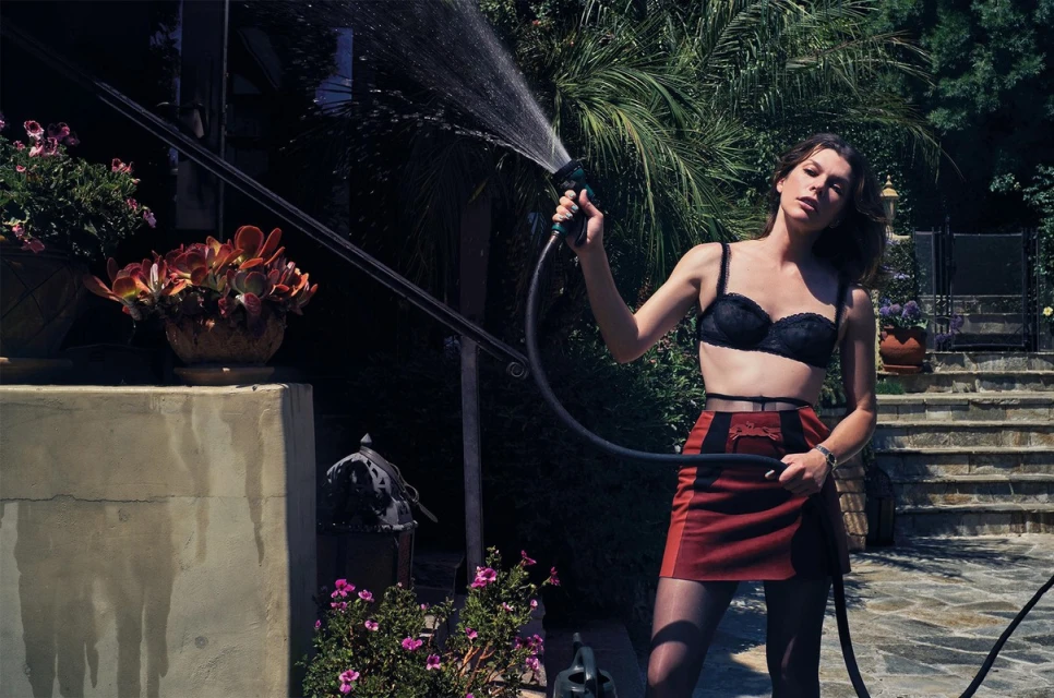 A young adult woman waters plants in a backyard garden wearing a black bra and rust-colored skirt with a water hose on a sunny day.