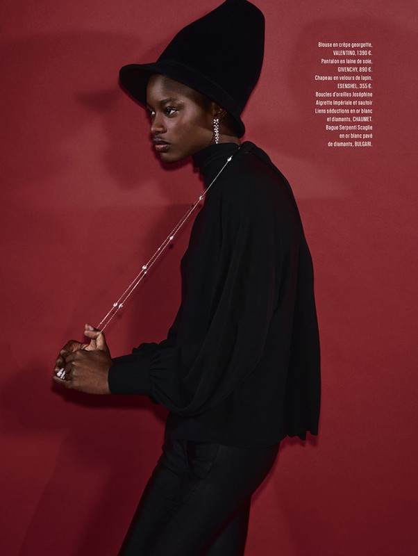 Fashion shot of a Black woman in a black turtleneck and hat, posing against a red background. She is wearing jewelry.