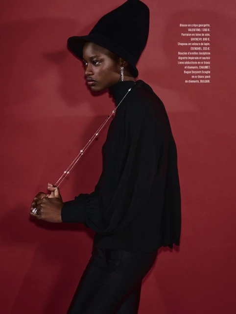 Fashion shot of a Black woman in a black turtleneck and hat, posing against a red background. She is wearing jewelry.