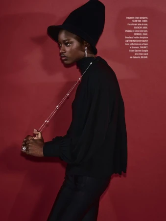 Fashion shot of a Black woman in a black turtleneck and hat, posing against a red background. She is wearing jewelry.